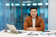 © Liubomir - Asian businessman seated at desk with laptop, documents nearby. Casual attire, genuine smile conveying professionalism, sincerity. Modern office setting reflecting successful corporate atmosphere.