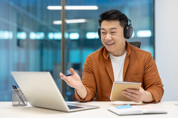 Wall Mural - Asian businessman wearing headphones engaging in virtual meeting, holding notebook. Smiling professional in office setting, using laptop for remote work and clear communication, showcasing modern