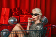 © k8most - Stylish senior woman with gray hair, wearing sunglasses and a silver dress, sits in a supermarket cart at a party, sipping wine against a backdrop of red curtains.