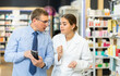 © JackF - Concerned middle-aged male customer asking young chemist about product reflected on his phone in large pharmacy