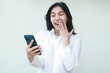 © M Alfan Setyawan - image of successful young woman asian manager surprised looking sale promo at smartphone covering open mouth with palms feeling shy wearing white suit shirt, using new apps, standing read good news