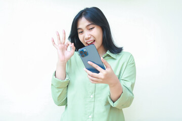 playful satisfied asian woman with dark hair wearing green over size shirt confident showing okay symbols looking at smartphone with joy recommend good product standing isolated white background