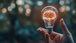 © Sinechana - 2410 79.A close-up of a hand presenting a virtual lightbulb with a glowing brain inside. The bokeh background gives a sense of energy and inspiration, emphasizing the concepts of smart thinking and
