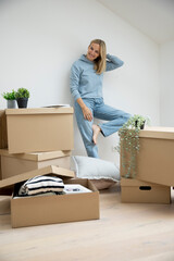 woman with blond hair stands in her new apartment, loft, house surrounded by many brown moving boxes