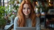 © Joachim - A cheerful red-haired woman uses her laptop in a serene, plant-filled environment, highlighting themes of connection, technology, and modern workspaces.