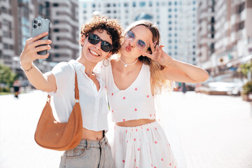 Two young beautiful smiling cute women in trendy summer sundress. Sexy carefree models posing in the street in sunny day. Positive models having fun and hugging. Cheerful and happy, take selfie