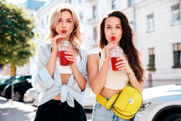 Two young beautiful smiling glam female in trendy summer clothes. Sexy women posing outdoors. Positive models holding and drinking fresh cocktail smoothie drink in plastic cup. Bright makeup, red lips