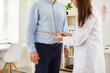 © Studio Romantic - Cropped photo of a young female doctor physiotherapist or nurse measure waist with measuring tape for male patient standing in medical clinic during examination. Weight loss and diet concept.