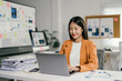 © PaeGAG - Young asian businesswoman is smiling while working on a laptop in a modern office, surrounded by financial charts and documents, reflecting a productive and positive work environment