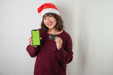 Excited young asian woman wearing santa clause hat and red long sleeved sweater is holding smartphone and credit card, isolated over white background. Concept for Christmas Holiday and New Year Party