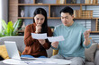 © Liubomir - Asian couple checking financial documents, looking concerned while discussing expenses at home. Man and woman focus on paperwork and stress about finances with laptop nearby.