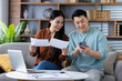 © Liubomir - Asian couple sitting on couch reviewing documents with laptop and phone. Smiling, discussing finances, teamwork, planning, comfort. Casual setting, teamwork in progress, happy atmosphere.