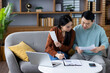 © Liubomir - Asian couple reviewing financial documents in living room, illustrating teamwork, home finance management. Man and woman using laptop, phone, and paperwork for planning, communication.