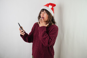 Excited young asian woman wearing santa clause hat and red long sleeved sweater is using smartphone, isolated over white background. Concept for Christmas Holiday and New Year Party