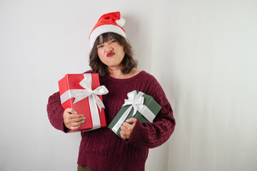 Excited young asian woman wearing santa clause hat and red long sleeved sweater is carrying large gift box with ribbon, isolated over white background. Concept for Christmas Holiday and New Year Party