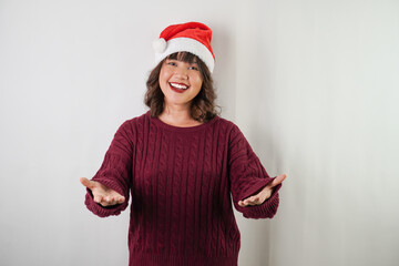 Friendly young asian woman wearing santa clause hat and red long sleeved sweater is showing empty opened hand palm, isolated over white background. Concept for Christmas Holiday and New Year Party