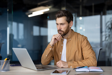 Mature businessman focused on laptop in office setting. Thoughtful expression indicates analysis and concentration. Notebook and phone on desk suggest professional work environment.