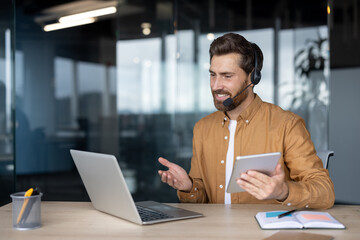 Mature businessman in headset conducts online meeting using laptop and tablet. Professional setup at office desk for effective remote communication. Engaged in digital discussion on business topics.