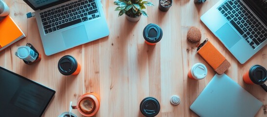 Top view of a wooden office desk with two laptops, coffee mugs, a plant, a phone, and a notebook.