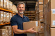 © abdlkerim - A middle-aged man is preparing to ship goods, smiling and holding cardboard boxes in the warehouse of his small business company for selling products online on an e-commerce platform.