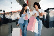 © LAUGH - Portrait of shopper girls walking outside of shopping mall with shopping bags in hands and looking at the phone while gossiping. Trendy shoppers have fun at shopping center and went for shopping spree