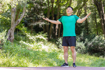 Sporty happy Japanese guy practicing yoga meditation, receives positive energy, healthy lifestyle. Young man standing making breath exercises, relaxing in park on sunny summer day. Active sportsman.