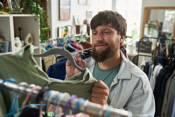 Young male shopper looking at secondhand shirt on hanger while choosing new items from collection of casual attire