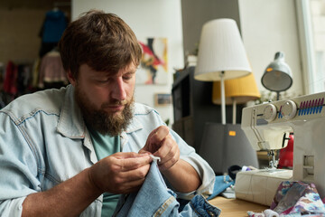 Young male worker of thrift shop sitting in front of camera and ripping blue secondhand jeans while upcycling used attire