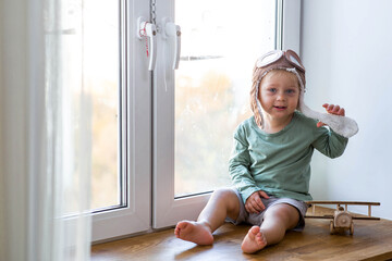 A cute 2-year-old boy in a cap is playing on the windowsill with a wooden airplane. Children's wooden toys for child development.