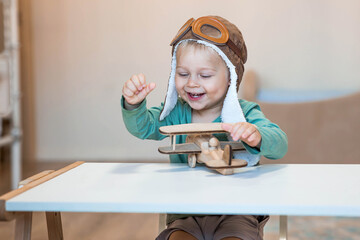 A handsome little boy of 2 years old in a pilot's hat is playing with a wooden airplane and a logistics constructor. Children's wooden toys for child development.