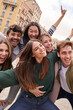 © CarlosBarquero - Vertical. Tourist group of young excited friends together posing happy for photo in city. Gen z people smiling with arms raised in air for holiday souvenir picture enjoying having fun in European town
