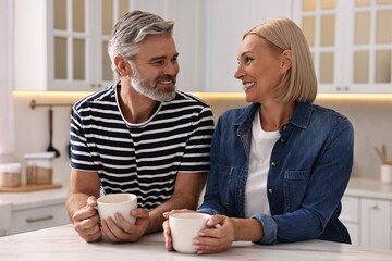 Wall Mural - Happy middle aged couple with cups of drink at white marble table in kitchen