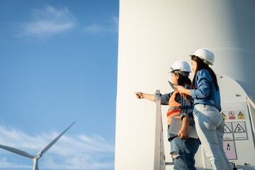 two engineers are  wearing hard hats and orange vests are pointing at a wind turbine. They are looking at the turbine and possibly discussing its design or operation