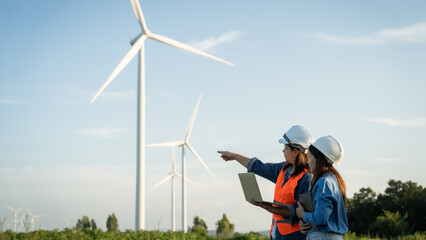 two engineers are standing in front of a wind turbine, one of them holding a laptop. Scene is professional and focused, as the two people are likely discussing the turbine's design or performance