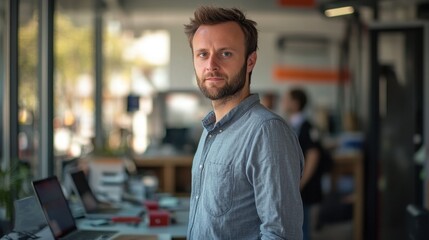 A young European man stands confidently in a tech startup. His thoughtful demeanor and casual attire reflect a modern workspace filled with innovation and collaboration.