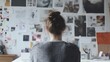 © Tcukimay - A woman with her hair in a bun sits at a desk facing a wall covered in photos and papers, seemingly lost in thought.