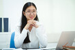 © NanSan - An Asian businesswoman smiles confidently while working at her desk in the office. She diligently analyzes financial data on her laptop, showcasing her expertise and commitment as an accountant