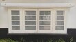 © Rifky Rachman Safri - A white-framed window with six glass panes stands out against a black, textured wall base. Reflecting the outdoors, the window is bordered by greenery, adding contrast to the scene.
