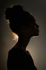 Silhouette of a young woman with an elegant bun hairstyle, softly illuminated against a dark background, conveying mystery and introspection.