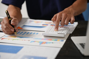 Wall Mural - A focused businessman sits at his office desk, analyzing a paper chart while working on his laptop. He examines financial and marketing reports, concentrating on data for strategic decision-making.