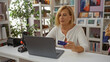 © Krakenimages.com - Mature woman with short blonde hair using a laptop in a decorated home office setting, holding a blue credit card, surrounded by plants and indoor decor items