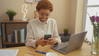 © Krakenimages.com - Smiling african woman using smartphone at home office with laptop, flowers, and modern decor