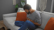© Krakenimages.com - Young man with a beard sitting on a couch in a living room, clutching his stomach in pain, wearing a striped shirt and jeans, surrounded by orange pillows and plants.
