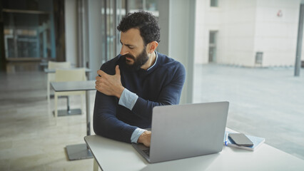 Middle-aged bearded man in pain holding shoulder while working on laptop in modern office.