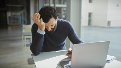 Stressed businessman with beard in modern office using laptop and holding head