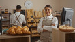 © Krakenimages.com - Woman smiling in bakery with arms crossed while man arranges bread in background, displaying various pastries and bread in indoor shop