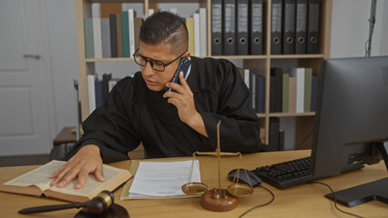 Young hispanic man judge at desk in an office reading book while talking on phone near computer and legal scales