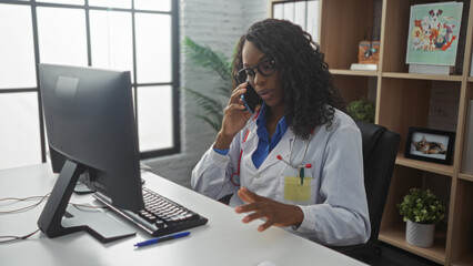 Wall Mural - A young african american woman doctor with curly hair talks on the phone in a modern clinic office while working on a computer.