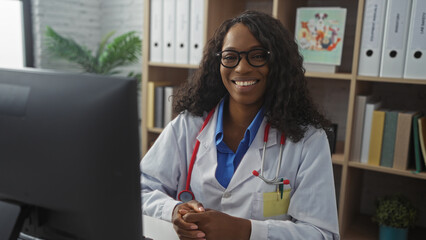 Wall Mural - Young african american woman doctor with curly hair and glasses in a hospital clinic room, wearing a lab coat and stethoscope, smiling confidently at her computer in an indoor workplace.