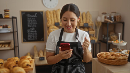 Young woman celebrating success with clenched fist while looking at her phone in a cozy bakery, surrounded by fresh bread and pastries.